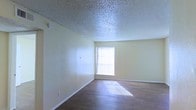 This single-story space presents a clean, open room characterized by warm hardwood-style flooring that runs throughout. The room is finished with smooth, light yellow walls that complement the natural light streaming in through a window dressed with vertical blinds. The textured popcorn ceiling adds a classic touch to the interior. White baseboards provide a crisp contrast against both the walls and flooring. The window serves as the room's primary light source, casting defined stripes of sunlight across the floor and creating a bright, airy atmosphere. The space is straightforward in layout, offering an open and versatile environment suitable for a variety of uses. Wall outlets are visibly placed for convenient access, and the overall condition of the room appears well-maintained. The minimalist design and neutral color palette make it an adaptable space that can accommodate different furniture arrangements and décor styles with ease.