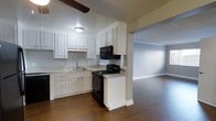 This single-floor residence offers a practical and well-appointed layout that seamlessly connects the kitchen and living areas. The kitchen is outfitted with crisp white raised-panel cabinetry that provides ample storage, complemented by dark black appliances including a range, microwave, and dishwasher. A double-basin sink sits beneath the cabinetry, and light-toned countertops offer functional prep space. A textured ceiling adds character overhead, while a ceiling fan aids in air circulation throughout the space. The kitchen opens directly into a spacious living area, creating an open, airy feel ideal for both everyday living and entertaining. Rich, dark hardwood flooring flows continuously from the kitchen through the living space, adding warmth and visual cohesion to the interior. Crown molding accents the walls, lending a refined architectural detail throughout. The living area is generously sized, offering flexibility for a variety of furniture arrangements. Neutral gray walls provide a versatile backdrop that works well with a range of décor styles. Overall, the space presents a clean, functional aesthetic with classic design details and a comfortable, connected floor plan.