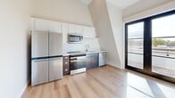 Located at 304 E Jackson Blvd in Elkhart, Indiana, this thoughtfully designed unit offers an efficient and modern living experience across a single floor. The open-concept layout seamlessly connects the living, dining, and kitchen areas, creating a functional and cohesive space. Soaring angled ceilings amplify the sense of openness, while large windows and a glass-panel door invite abundant natural light and provide outdoor views.

The kitchen is well-appointed with stainless steel appliances — including a refrigerator, range, microwave, and dishwasher — complemented by two-tone cabinetry featuring white upper cabinets and darker lower units, a classic white subway tile backsplash, and ample counter space. Light hardwood-style flooring runs throughout the entire unit, lending warmth and continuity to every room.

The living area is bright and open, with clean white walls and recessed lighting that enhance the airy atmosphere. The entry features a stylish white paneled door with modern black hardware, framed by sun-drenched floors.

The bathroom is clean and contemporary, finished with white walls and the same light hardwood flooring. It includes a full shower-tub combination, a toilet, and a stacked washer and dryer unit, adding notable convenience to daily living.