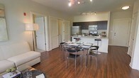 This single-floor residence showcases a thoughtfully designed open-concept layout that connects the living, dining, and kitchen areas into one cohesive and functional space. Rich, dark hardwood flooring runs throughout, adding warmth and a polished aesthetic to the interior.

The kitchen is outfitted with dark espresso cabinetry, stainless steel appliances including a refrigerator, microwave, and range, and a light-toned countertop that provides a striking contrast to the cabinetry. A breakfast bar with modern bar stools offers a casual seating option, while the overall layout provides ample workspace for cooking and entertaining.

Adjacent to the kitchen, the dining area accommodates a sleek glass-top table with contemporary chrome-framed chairs, ideal for both everyday meals and hosting guests. Track lighting overhead ensures the entire open space is well-lit and highlights the modern design elements throughout.

The living area flows naturally from the dining space, offering a relaxed and welcoming atmosphere. Several interior doors are visible, including a louvered closet door, suggesting convenient storage solutions within the residence. The overall ambiance is modern, clean, and functional, with a cohesive design language carried throughout the space.