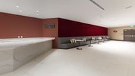 Panorama Tower's private cinema amenity is a thoughtfully designed entertainment space that seamlessly blends a sophisticated lounge with a full-scale screening room. The lounge area sets a refined tone with its expansive white marble-veined flooring, recessed lighting, and a striking marble bar counter lined with sleek metal barstools. Deep crimson accent walls — including a bold upholstered panel — add dramatic color and texture, while a long banquette of charcoal leather seating paired with pedestal cocktail tables creates an inviting pre- or post-screening gathering area. Film-themed décor and a wall-mounted TV complete the lounge's cinematic ambiance. Transitioning into the screening room, the space shifts to an immersive, darkened environment with matte black walls and ceilings that eliminate distraction. Tiered rows of red-and-black upholstered theater seats with built-in cup holders face a large projection screen, ensuring excellent sightlines from every position. Textured gray carpet absorbs sound and enhances acoustics throughout the room. Together, the lounge and screening room form a cohesive, premium cinema experience well-suited for private events, movie nights, and curated screenings within Panorama Tower.