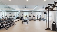Located at 1830 Autumn Dr in Hoffman Estates, IL, this fitness center offers a generously sized, single-floor layout designed to accommodate a comprehensive range of workout activities. The space is divided into distinct functional zones, each outfitted with purpose-specific equipment.

The cardio area features multiple treadmills, elliptical trainers, stair climbers, and a stationary bike, all positioned near large windows that allow abundant natural light and views of the surrounding exterior. A full-length wall mirror visually expands the space and supports proper form during exercise.

The strength and free weights zone includes a structured dumbbell rack with a wide weight range, adjustable benches, and open floor space for functional movements. A dedicated functional training area houses a multi-station cable machine rig, TRX suspension trainers, a wall-mounted storage system for resistance accessories, and specialty equipment such as a tire sled for high-intensity training.

Overhead, exposed ductwork and pendant linear lighting fixtures contribute to an industrial-modern aesthetic, while ceiling fans promote air circulation throughout. The durable speckled rubber flooring provides both safety and comfort underfoot. The overall design is clean, bright, and organized, creating an efficient and motivating environment for fitness.