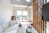 This thoughtfully designed modern unit offers a striking blend of contemporary architecture and warm, natural materials. The open-plan living area commands attention with soaring double-height ceilings and clerestory windows that flood the space with abundant natural light, complemented by large sliding glass doors that frame outdoor views and further brighten the interior. Light-toned hardwood flooring runs throughout, lending a cohesive warmth to every room.

A vertical wood slat partition elegantly separates the living area from the staircase, adding texture and visual interest while maintaining an airy, open feel. The bold cobalt-blue staircase, accented with a natural wood handrail, serves as a striking architectural focal point, leading up to a lofted sleeping area. The bedroom loft is a serene retreat, featuring clean white walls, a ceiling fan, and a low-profile platform bed that enhances the minimalist aesthetic.

The galley-style kitchen is efficiently appointed with warm wood cabinetry, white tile backsplash, stainless steel appliances — including a full-size refrigerator, range, dishwasher, and built-in microwave — and ample counter space. Track lighting throughout the unit provides flexible, modern illumination. Overall, the space exudes a clean, minimalist ambiance with thoughtful design details at every turn.
