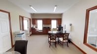 This single-story professional office space combines functionality with a warm, polished aesthetic. The layout includes a reception and meeting area, a private office, and supporting workspaces, all finished with neutral tones, carpeted floors, and wood trim accents that lend a cohesive, professional feel throughout.

The reception and client seating area serves as the central gathering space, featuring a white fireplace mantle that adds a welcoming residential touch. A round glass-top table surrounded by dark leather chairs provides an inviting consultation setting. Framed informational displays and brochure holders are neatly arranged, indicating active client-facing use. Glass-paned French doors connect this area to an adjacent tiled space, enhancing the sense of openness and flow.

The private office is notably well-appointed, featuring floor-to-ceiling rich mahogany cabinetry with decorative crown detailing and dark countertops running the length of one wall. Two windows with blinds allow natural light into the space. A glass-top executive desk with upholstered guest chairs anchors the room, while built-in workspace and office equipment are neatly integrated into the cabinetry. Fluorescent strip lighting provides consistent illumination throughout, and a security camera is visible, reflecting practical operational considerations.