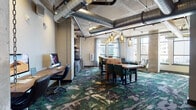 This single-floor space showcases a compelling industrial-modern aesthetic, blending raw architectural elements with carefully curated furnishings. Exposed silver ductwork runs prominently across the ceiling alongside concrete columns, establishing an urban, loft-like character throughout. Large windows line the perimeter, flooding the interior with generous natural light and offering views of the surrounding cityscape.

The main area centers around substantial live-edge wooden communal tables paired with emerald green upholstered chairs, illuminated overhead by clusters of amber glass pendant lights. A bold, abstract-patterned carpet in deep greens, grays, and earth tones anchors the space and adds a distinctive visual element. A dedicated computer workstation area features a wall-mounted live-edge wood desk with multiple desktop computers, suspended by industrial chains — a functional yet stylized touch.

The kitchenette is a standout feature, finished with reclaimed wood cabinetry, a dark stone countertop, glossy black subway tile backsplash, open wood shelving, a built-in oven, and a beverage refrigerator. Exposed brick accent walls add warmth and texture. A private room with a frosted-glass door and wall-mounted display provides an enclosed meeting option. Thoughtful artwork, including a dramatic train-themed piece, completes the space's industrial narrative.