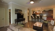 This single-story space showcases an open-concept layout that seamlessly connects the living area to the kitchen, creating a cohesive and functional environment. The living area features dark hardwood flooring, crown molding, and a ceiling fan with a light fixture, contributing to a well-lit and comfortable atmosphere. A media console with a large flat-screen television anchors one wall, accompanied by a decorative stacked-stone wall accent that adds texture and visual interest. A plush sofa and a dark wood coffee table complete the seating area. The kitchen is visible from the living space and is equipped with warm wood cabinetry, stainless steel appliances, and a tile backsplash. A kitchen island with a raised bar counter accommodates multiple bar-height chairs, making it an ideal spot for casual dining or entertaining. Pendant lights hang above the island, providing focused illumination and enhancing the warm ambiance of the space. The open sightlines between the two areas make the overall layout feel expansive and inviting, while the consistent use of dark wood tones and neutral wall colors creates a cohesive design aesthetic throughout.