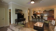 This single-story space showcases an open-concept layout that seamlessly connects the living area to the kitchen, creating a cohesive and functional environment. The living area features dark hardwood flooring, crown molding, and a ceiling fan with a light fixture, contributing to a well-lit and comfortable atmosphere. A media console with a large flat-screen television anchors one wall, accompanied by a decorative stacked-stone wall accent that adds texture and visual interest. A plush sofa and a dark wood coffee table complete the seating area. The kitchen is visible from the living space and is equipped with warm wood cabinetry, stainless steel appliances, and a tile backsplash. A kitchen island with a raised bar counter accommodates multiple bar-height chairs, making it an ideal spot for casual dining or entertaining. Pendant lights hang above the island, providing focused illumination and enhancing the warm ambiance of the space. The open sightlines between the two areas make the overall layout feel expansive and inviting, while the consistent use of dark wood tones and neutral wall colors creates a cohesive design aesthetic throughout.