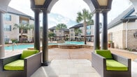 Located at 3918 Atascocita Rd in Humble, Texas, this expansive outdoor amenity area showcases a resort-inspired design that serves as a central gathering space for the community. The centerpiece is a large, freeform swimming pool with gently curving edges and crystal-clear blue water, complemented by a raised spa feature finished in warm stone tile. Tall, swaying palm trees are strategically placed throughout the space, evoking a tropical atmosphere and providing natural shade across the stamped concrete sundeck. A striking clubhouse structure anchors one side of the pool area, featuring a distinctive Tudor-inspired gabled roofline, arched colonnades with bold columns, and stucco and brick exterior detailing. A separate covered pergola cabana offers a shaded lounge area furnished with comfortable seating, ideal for relaxing poolside. The surrounding multi-story residential buildings feature classic brick and stone exteriors with dark rooflines and private balconies overlooking the pool courtyard. Outdoor dining sets and lounge chairs are distributed across the deck, providing ample space for both social gatherings and quiet relaxation. Lush, manicured landscaping borders the entire area, enhancing the clean, well-appointed aesthetic of this outdoor retreat.