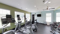 This fitness center offers a practical and welcoming environment for exercise. The space is finished with light-colored walls in soft green and blue tones, complemented by a white ceiling with recessed lighting and a ceiling fan that helps maintain a comfortable atmosphere during workouts. Dark, durable flooring lines the room, providing a resilient surface suited for active use. Multiple windows dressed with white blinds allow natural light to fill the space while offering views of the surrounding greenery outside. The cardio area features several elliptical machines, each equipped with individual monitors for personalized entertainment during workouts. A multi-station strength training machine anchors the equipment selection, providing options for a variety of resistance exercises. A wall-mounted flat-screen television is positioned for easy viewing from multiple workout stations. A small rack near the wall offers additional accessories or towels for convenience. The overall layout is organized and functional, allowing users to move between equipment comfortably. The combination of natural light, ventilation, and varied equipment makes this fitness center a well-rounded facility for maintaining an active lifestyle.