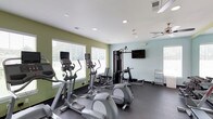 This fitness center offers a practical and welcoming environment for exercise. The space is finished with light-colored walls in soft green and blue tones, complemented by a white ceiling with recessed lighting and a ceiling fan that helps maintain a comfortable atmosphere during workouts. Dark, durable flooring lines the room, providing a resilient surface suited for active use. Multiple windows dressed with white blinds allow natural light to fill the space while offering views of the surrounding greenery outside. The cardio area features several elliptical machines, each equipped with individual monitors for personalized entertainment during workouts. A multi-station strength training machine anchors the equipment selection, providing options for a variety of resistance exercises. A wall-mounted flat-screen television is positioned for easy viewing from multiple workout stations. A small rack near the wall offers additional accessories or towels for convenience. The overall layout is organized and functional, allowing users to move between equipment comfortably. The combination of natural light, ventilation, and varied equipment makes this fitness center a well-rounded facility for maintaining an active lifestyle.