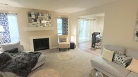This single-floor residence offers a thoughtfully designed open-concept layout that seamlessly connects the living and dining areas, creating a spacious and inviting atmosphere. The living room is anchored by a classic white fireplace, above which a striking geometric mirrored wall art piece serves as a bold focal point. Soft, neutral wall tones and plush carpeting throughout lend the space a warm and cohesive aesthetic. Large windows dressed with patterned and sheer curtains fill the interior with generous natural light, enhancing the airy feel of the room. The living area is furnished with contemporary grey sofas, layered with circular shag rugs that add texture and depth to the flooring. A chic accent chair and side lamp provide an intimate reading nook near the window. Through an open archway, the dining area is visible, featuring a sleek dark dining set with glass accents, complemented by light sheer curtains that continue the bright, elegant theme. The overall ambiance of the space is modern yet comfortable, blending clean lines with warm, neutral finishes to create a home that feels both polished and welcoming.