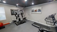 This dedicated fitness space is designed to support a complete workout experience within a single, efficiently organized room. The space features a multi-station cable gym unit positioned atop a protective rubber mat, offering a wide range of strength-training exercises. A Vision Fitness treadmill provides cardio capability, rounding out the exercise options available. Wall-mounted mirrors along one side enhance the sense of space and allow users to monitor their form during workouts. The room is finished with neutral gray walls and matching gray carpet, creating a clean and focused environment. A striking triptych artwork depicting classic vintage automobiles adds a personalized decorative touch, bringing character to the functional space. The dark-painted ceiling, accented by recessed lighting, lends a modern and polished aesthetic that distinguishes this fitness room from a conventional setup. Crown molding along the upper walls adds a refined architectural detail. The recessed lighting provides bright, even illumination throughout the space, ensuring excellent visibility during any type of exercise. Overall, this fitness room balances practicality and style, making it a well-appointed space for daily training.