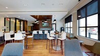 This contemporary single-floor communal space is thoughtfully designed to blend functionality with modern aesthetics. The open-plan layout is anchored by warm hardwood flooring that runs throughout, creating a cohesive and inviting atmosphere. At the heart of the space is a sleek bar-style counter, framed by a striking wood-paneled accent wall and ceiling detail that draws the eye upward and adds architectural depth. A wall-mounted flat-screen display is integrated into this focal feature, making the area well-suited for social gatherings or casual viewing. Pendant lighting in matte black hangs above the central zone, contributing a refined, modern edge to the overall design. Multiple high-top bistro tables paired with contemporary white and chrome bar stools are distributed throughout, offering flexible seating arrangements for individuals or small groups. Toward the perimeter, large black-framed windows invite abundant natural light, complemented by a cozy lounge seating area with upholstered sofas and chairs. Recessed lighting throughout the ceiling ensures the space remains well-lit at all times. A beverage station is also visible, enhancing the space's utility as a social hub. The overall ambiance is polished, welcoming, and well-appointed for modern communal use.