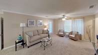 Barrington 22 is a thoughtfully designed single-floor residence that offers a comfortable and cohesive living experience. The space is finished in warm neutral tones throughout, creating a welcoming ambiance from room to room. The living area is generously sized, featuring plush carpeting, crown molding, a ceiling fan, and ample natural light streaming through multiple windows dressed with sheer curtains. A private exterior door in the living space adds convenient access. The adjoining dining area is defined by an elegant pendant light fixture and crown molding, providing an intimate setting for meals. The kitchen is efficiently laid out with white cabinetry, a built-in wine rack, wood-look flooring, and black appliances including a range and refrigerator. Multiple bedrooms offer carpeted floors, ceiling fans, and natural light through curtained windows, each providing a restful retreat. The primary bedroom includes a wall-mounted TV and a wicker accent chair. The primary bathroom features a wide vanity, expansive mirror, wood-look flooring, and generous counter space. A second full bathroom offers a tub-shower combination, single vanity, and matching warm-toned finishes. Together, these spaces form a complete and functional home with consistent, understated style.
