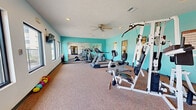 Located at 1001 Tramway Blvd NE in Albuquerque, New Mexico, this fitness center offers a well-organized and functional workout space on a single floor. The room is finished with wall-to-wall carpeting and features a textured ceiling with recessed lighting and a ceiling fan, keeping the environment comfortable during workouts. Vibrant teal accent walls add a lively, energetic atmosphere to the space, complemented by crisp white walls on the opposing sides.

The facility is equipped with a variety of exercise machines, including treadmills, a stationary bike, a multi-station cable and weight machine, and a rowing-style machine, providing options for both cardiovascular and strength training. A set of colorful kettlebells is also available for free-weight exercises. A large wall-mounted flat-screen television is positioned for easy viewing during workouts.

Full-length mirrors line one wall, visually expanding the space and allowing users to monitor their form. Large windows along the perimeter flood the room with natural light and offer direct views of an outdoor pool area, creating a refreshing connection to the outdoors. Glass double doors provide access to adjacent amenity spaces. The overall ambiance is bright, clean, and purpose-driven, making it a practical and inviting facility for residents.