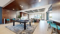 Located at 4400 Washington Street in Kansas City, MO, this single-floor amenity space combines contemporary design with upscale finishes to create a refined communal environment. The entry foyer welcomes visitors with floor-to-ceiling glass doors, large-format tile flooring, and abundant natural light streaming in from the surrounding courtyard views.

Moving inward, the lobby lounge is defined by a coffered ceiling with subtle ambient lighting, rich wood panel accents, and bold artwork depicting Kansas City landmarks. Curated seating arrangements—featuring plush armchairs in teal, yellow, and neutral tones—are anchored by layered area rugs, lending a welcoming residential character to the space.

Adjacent to the lobby, a spacious game and entertainment lounge features a full-size billiards table positioned atop an abstract area rug. This area transitions seamlessly into a fireplace lounge with marble surround and a wall-mounted television, flanked by a bar counter with teal stools. A coffered tray ceiling with warm wood paneling defines the zone architecturally.

The fully equipped catering kitchen showcases a dramatic waterfall-edge marble island with leather bar seating, high-gloss cabinetry, stainless steel appliances, and a geometric tile backsplash. Floor-to-ceiling windows bathe the kitchen in natural light while offering outdoor views.