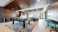Located at 4400 Washington Street in Kansas City, MO, this single-floor amenity space combines contemporary design with upscale finishes to create a refined communal environment. The entry foyer welcomes visitors with floor-to-ceiling glass doors, large-format tile flooring, and abundant natural light streaming in from the surrounding courtyard views.

Moving inward, the lobby lounge is defined by a coffered ceiling with subtle ambient lighting, rich wood panel accents, and bold artwork depicting Kansas City landmarks. Curated seating arrangements—featuring plush armchairs in teal, yellow, and neutral tones—are anchored by layered area rugs, lending a welcoming residential character to the space.

Adjacent to the lobby, a spacious game and entertainment lounge features a full-size billiards table positioned atop an abstract area rug. This area transitions seamlessly into a fireplace lounge with marble surround and a wall-mounted television, flanked by a bar counter with teal stools. A coffered tray ceiling with warm wood paneling defines the zone architecturally.

The fully equipped catering kitchen showcases a dramatic waterfall-edge marble island with leather bar seating, high-gloss cabinetry, stainless steel appliances, and a geometric tile backsplash. Floor-to-ceiling windows bathe the kitchen in natural light while offering outdoor views.