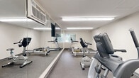 This dedicated fitness facility is a single-room exercise space designed to maximize functionality within a compact footprint. The room features durable speckled rubber flooring throughout, providing a safe and resilient surface suited for active use. Bright fluorescent lighting illuminates the space evenly, ensuring excellent visibility during workouts at any time of day.

A large full-wall mirror runs along one side of the room, visually expanding the space and allowing users to monitor their form. A wall-mounted flat-screen TV is positioned prominently above the mirror, offering entertainment during exercise sessions. A wall-mounted HVAC unit provides climate control for year-round comfort.

The room is outfitted with a well-rounded selection of cardio equipment, including a treadmill, elliptical trainer, stationary bike, rowing machine, and a stair climber. A window along one wall allows natural light to filter in, offering a view of the surrounding greenery. A set of glass-panel doors provides direct access to an outdoor area, enhancing ventilation and connection to the exterior.

Practical amenities include a small bench and shoe rack near the entrance, a sanitation station with wipes and supplies for equipment cleaning, wall-mounted shelving for personal items, and a security camera for added safety. Posted guidelines and framed informational documents keep the space organized and user-friendly.
