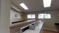 This single-floor laundry facility is designed for efficiency and practicality, offering residents a clean and functional space to handle their laundry needs. The room features a row of top-loading washing machines aligned along a continuous counter surface, providing ample workspace for sorting and folding clothes. A generous built-in countertop runs along the opposite wall, offering additional space for organizing laundry items.

Natural light fills the room through a series of horizontal windows positioned along the upper portion of the walls, offering views of the surrounding greenery outside. The space is further illuminated by recessed fluorescent ceiling panels, ensuring consistently bright conditions throughout the day and into the evening.

The walls are finished in a neutral warm beige tone, contributing to a clean and inviting atmosphere. Decorative framed artwork adds a subtle personal touch to the otherwise utilitarian environment. The flooring is composed of light hardwood, adding warmth to the space. Ventilation is provided through ceiling-mounted vents, maintaining comfortable air quality. Overall, the facility is thoughtfully laid out to maximize usability while maintaining a tidy and welcoming environment.