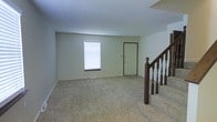 This two-story residence offers a functional and practical layout spread across two floors. The main level features a spacious carpeted living area that provides a warm and comfortable foundation for the space. Natural light filters in through a window fitted with horizontal blinds, brightening the room with a soft, neutral ambiance. The light-toned walls and beige carpet create a clean, understated aesthetic throughout.

A classic wood staircase with dark-stained balusters and a prominent newel post serves as a central architectural feature, connecting the main level to the upper floor. The staircase adds a touch of traditional character to the otherwise simple interior. A door on the main level provides additional access or utility, contributing to the overall practicality of the floor plan.

The space is finished with standard trim and baseboards that frame each room neatly. The carpet, consistent throughout the visible areas, adds softness underfoot and contributes to the residential warmth of the home. Overall, the space presents a straightforward, move-in-ready interior with a neutral palette that offers flexibility for a variety of furnishing and décor styles across both floors.
