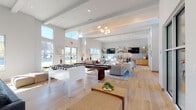 This thoughtfully designed single-floor amenity space offers a expansive, open-concept layout bathed in natural light streaming through floor-to-ceiling windows and clerestory glazing. The soaring ceiling height, accented by exposed beam detailing and contemporary pendant chandeliers, creates an airy, welcoming atmosphere throughout the main lounge area, which features light wood-look flooring, plush seating arrangements, and game amenities including a foosball table and billiards table.

A dedicated dining area showcases a substantial wood communal table surrounded by cane-back chairs beneath a row of oversized circular brass pendant lights. Open wood shelving lines the walls, styled with decorative objects and dual wall-mounted televisions, while large glass doors open directly onto an outdoor patio featuring grilling stations and al fresco seating.

A separate lounge nook provides an intimate retreat, distinguished by a striking textured stone accent wall hosting a wall-mounted television, accompanied by a built-in wet bar with stainless steel appliances and casual seating. An additional cozy sitting area features warm cognac leather sofas, globe pendant lighting, and curated wall art, completing a multi-functional space designed for both social gatherings and relaxed leisure.
