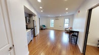 This single-floor space offers an open-plan layout that seamlessly integrates the kitchen and living areas into one expansive, versatile environment. The warm hardwood flooring extends throughout, adding a sense of continuity and richness to the interior. Recessed ceiling lights are distributed evenly across the white ceiling, providing consistent, bright illumination throughout the space.

The kitchen is positioned along one wall and features gray cabinetry, stainless steel appliances including a refrigerator, microwave, and range, along with a clean white countertop. It is partially separated from the main living area by a low partition, maintaining an open feel while defining its own functional zone.

The living area is generously proportioned, offering ample room for various furniture arrangements. Multiple windows along the exterior wall allow natural light to fill the space, contributing to its airy atmosphere. A wall-mounted split air conditioning unit provides climate control. The white walls and clean architectural lines give the space a modern, neutral aesthetic, making it highly adaptable to a range of uses.