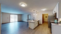 This single-floor residence offers a thoughtfully designed layout that balances open communal spaces with well-defined private areas. The heart of the home is an open-concept kitchen and living area, where durable wood-look flooring flows seamlessly underfoot. The kitchen is outfitted with crisp white cabinetry, black hardware accents, light quartz countertops, and a full suite of stainless steel appliances including a refrigerator, range, microwave, and dishwasher. A central island with bar seating serves as both a prep space and casual dining area, illuminated by stylish pendant lights. The adjacent living area is spacious and bright, with sliding glass doors that invite natural light and provide outdoor access. Bedrooms feature plush carpeting, warm wood-trimmed windows, and generous proportions. The bathrooms are clean and modern, with white vanities, vessel or undermount sinks, black hardware, and wood-look flooring. A dedicated hallway connects the private quarters efficiently. A functional laundry room houses a full-size washer and dryer. Warm wood door and trim details are consistent throughout, lending a cohesive, polished character to the entire residence.