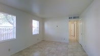 This two-floor space offers a bright and clean interior defined by its simple, functional design. The living area is finished with light-toned square tile flooring that extends seamlessly throughout, contributing to a cohesive and airy feel. White walls and smooth ceilings reflect natural light effectively, keeping the interior feeling open and well-lit. A window fitted with blinds allows natural light to filter into the main room while maintaining privacy. Ceiling-mounted HVAC vents are integrated into the layout, ensuring efficient climate control throughout. The neutral color palette and minimal architectural detailing create a versatile backdrop suitable for a variety of interior styles. A hallway visible from the main living area leads to additional rooms, suggesting a thoughtful floor plan with clear circulation between spaces. A door at the end of the corridor provides access to further areas of the home, hinting at a well-organized spatial arrangement across both levels. Overall, this space presents a tidy, move-in ready environment with a clean aesthetic and practical features well-suited for daily living.