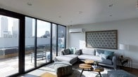 This contemporary single-floor space showcases a sleek, modern design that emphasizes natural light and urban connectivity. The living area is anchored by large floor-to-ceiling sliding glass doors with black-framed detailing, flooding the interior with daylight and offering direct access to a private outdoor terrace overlooking a vibrant cityscape. The terrace provides an elevated outdoor retreat complete with seating, ideal for taking in expansive views of the surrounding skyline.

Inside, the living room is thoughtfully appointed with a generous L-shaped sectional sofa in soft grey tones, complemented by teal and patterned accent cushions that add a touch of color. A pair of round, natural wood coffee tables ground the seating area, while a decorative laser-cut wall panel in a geometric star pattern serves as a striking focal feature. Light-toned flooring runs throughout, enhancing the bright and airy atmosphere.

Climate control is provided by a wall-mounted split-system air conditioning unit, ensuring year-round comfort. Recessed ceiling lighting and clean white walls contribute to the space's minimalist aesthetic. Overall, the design balances contemporary style with functional living, creating a refined and welcoming environment with strong urban character.
