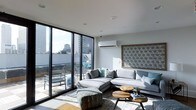 This contemporary single-floor space showcases a sleek, modern design that emphasizes natural light and urban connectivity. The living area is anchored by large floor-to-ceiling sliding glass doors with black-framed detailing, flooding the interior with daylight and offering direct access to a private outdoor terrace overlooking a vibrant cityscape. The terrace provides an elevated outdoor retreat complete with seating, ideal for taking in expansive views of the surrounding skyline.

Inside, the living room is thoughtfully appointed with a generous L-shaped sectional sofa in soft grey tones, complemented by teal and patterned accent cushions that add a touch of color. A pair of round, natural wood coffee tables ground the seating area, while a decorative laser-cut wall panel in a geometric star pattern serves as a striking focal feature. Light-toned flooring runs throughout, enhancing the bright and airy atmosphere.

Climate control is provided by a wall-mounted split-system air conditioning unit, ensuring year-round comfort. Recessed ceiling lighting and clean white walls contribute to the space's minimalist aesthetic. Overall, the design balances contemporary style with functional living, creating a refined and welcoming environment with strong urban character.