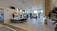 This sophisticated lobby space makes a striking first impression with its soaring ceilings and open, airy layout. The design centers around a sleek, contemporary reception desk finished in warm wood tones and topped with a white surface, accented by a series of gold cylindrical pendant lights and a dramatic spherical crystal chandelier overhead. Recessed lighting throughout the ceiling further enhances the bright, welcoming atmosphere.

Large floor-to-ceiling windows wrap around the space, flooding the interior with abundant natural light and offering clear views to the outdoors. The flooring features large-format neutral tiles, lending a clean and polished aesthetic that complements the overall modern design.

A comfortable lounge seating area is positioned near the windows, furnished with upholstered chairs and a coffee table, creating an inviting space for guests to relax. Tasteful decorative touches — including colorful abstract artwork, fresh floral arrangements, and potted plants — add warmth and visual interest throughout.

A textured white accent wall provides an architectural focal point, while the carefully curated furnishings and finishes come together to create a professional yet welcoming environment that balances elegance with functionality.