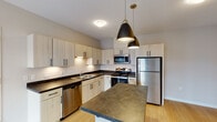 Located at 1350 Prairie Pkwy SW in Wyoming, Michigan, this single-floor unit offers a thoughtfully designed layout with modern finishes throughout. The kitchen serves as a central highlight, showcasing light-toned cabinetry with sleek black hardware, dark stone-style countertops, and a full suite of stainless steel appliances including a range, over-the-range microwave, refrigerator, and dishwasher. A central island with matching countertop provides additional prep space and casual seating, while stylish pendant lighting adds a contemporary touch overhead. Light wood-style flooring runs throughout the kitchen and living areas, contributing to a warm, cohesive aesthetic. A stacked washer and dryer unit is conveniently accessible just off the main living space, adding in-unit laundry functionality. The bedroom features neutral-toned carpet, soft wall colors, and a window with blinds that invites natural light into the space. The bathroom is clean and functional, featuring a full soaking tub with shower, a vanity with a light countertop, brushed nickel hardware accents, and dark tile flooring that grounds the overall neutral palette. The unit presents a modern, move-in-ready atmosphere well-suited for everyday living.
