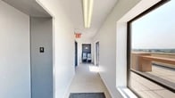 Located at 26-38 21st Street in Astoria, Queens, this rooftop amenity space offers an exceptional outdoor experience with sweeping, panoramic views of the New York City skyline, the Triborough (RFK) Bridge, and surrounding boroughs. The space is accessed via a bright, carpeted interior corridor with clean white walls, overhead fluorescent lighting, and a dedicated seating area near the rooftop entrance.

The expansive rooftop terrace is laid with large-format concrete pavers and is organized into several distinct zones. A sophisticated lounge area features contemporary white upholstered sofas and chairs arranged around a concrete coffee table, ideal for relaxed gatherings. A separate seating zone offers Adirondack-style chairs and rustic wood-framed furniture with cushioned seating. A row of teal-cushioned sun loungers provides ample space for sunbathing, positioned to take full advantage of the open sky. A charming wooden pergola structure serves as a covered bar area, complemented by warm wood-slatted built-in bench seating and planter boxes with greenery. The rooftop's elevated position delivers unobstructed 360-degree views, including the Manhattan and Queens skylines, making it a standout communal outdoor destination.