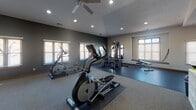 This fitness center is thoughtfully designed to accommodate a full range of workout needs across a single-floor layout. The main workout area is generously sized, featuring vaulted ceilings with dual ceiling fans and recessed lighting that keep the space bright and comfortable. Dark accent walls contrast with light neutral tones, creating an energizing atmosphere. Multiple windows fitted with wood-slat blinds allow natural light to fill the room while maintaining privacy.

The cardio section is well-stocked with True-brand treadmills, upright and recumbent bikes, and several elliptical trainers, all placed on protective rubber mats. A large wall-mounted mirror runs along one wall, accompanied by a row of flat-screen TVs, making cardio sessions more engaging. A water fountain is conveniently accessible nearby.

A dedicated strength-training zone houses a multi-station cable machine and a weight bench, while a separate free weights area provides a full dumbbell rack alongside stability balls for functional training. Wall-mounted coat hooks, a small storage unit, and a waste bin keep this section tidy and practical.

An adjoining stretching and floor exercise room offers a quieter environment, complete with padded floor mats and ample natural light from double windows. The carpeted and rubber-floored surfaces throughout provide comfort and traction, making this fitness center a comprehensive and inviting space for daily exercise.