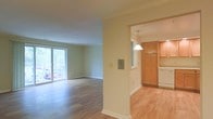 This single-floor space offers a clean, open layout finished with hardwood flooring throughout. The living area is generously proportioned, benefiting from abundant natural light that streams in through a large sliding glass door, which also provides views of a lush, tree-lined outdoor setting. Crown molding trims the ceilings, adding a refined architectural detail that runs consistently across the space.

A structural column visually separates the living area from the adjoining kitchen while maintaining an open, connected feel. The kitchen features warm honey-toned wood cabinetry extending to the upper walls for ample storage, light wood-toned flooring, and a white appliance finish. A pendant light with a classic dome shade hangs overhead, providing focused task lighting. A pass-through or window element connects the kitchen to an additional adjoining area, enhancing the sense of openness.

The walls are finished in a soft, neutral cream tone throughout, contributing to a bright and airy ambiance. White baseboards and trim provide crisp contrast, completing the overall clean and well-maintained character of the space.