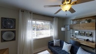 Located at 5802 University in San Diego, this single-floor residence offers a comfortable and well-appointed living environment. The living area serves as the heart of the home, showcasing a bright and airy atmosphere enhanced by a large window dressed with layered sheer curtains and horizontal blinds that allow abundant natural light to fill the room. A ceiling fan with an integrated light fixture anchors the space from above, providing both comfort and functionality. The living area is thoughtfully arranged, featuring a tufted navy blue sofa adorned with accent pillows, and is complemented by warm hardwood-style flooring that adds a sense of warmth and continuity throughout. An open shelving unit along the wall provides both display and storage options, reflecting a modern yet casual aesthetic. The neutral wall tones and carefully chosen furnishings create a cohesive, inviting ambiance suited for everyday living. The home's single-floor layout ensures ease of movement and accessibility, making it a practical and stylish residence in the vibrant San Diego community.