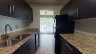 Located at 1346 Carriage Hill Ln in Hamilton, Ohio, this single-floor residence offers a well-organized layout with distinct living, sleeping, kitchen, and bathroom areas. The kitchen is a standout feature, designed in a galley style with dark espresso cabinetry, granite countertops, a stainless steel sink, a dishwasher, and a black refrigerator. Light wood-style flooring runs throughout the kitchen, which opens directly toward a living area bathed in natural light from large sliding glass doors fitted with vertical blinds. The living area transitions between wood-style and carpeted flooring, providing a comfortable and inviting atmosphere with views of surrounding greenery. The bedrooms are generously sized, each finished with neutral-toned carpet, textured ceilings, and windows that allow natural light to filter in. The bathroom is clean and functional, featuring a granite-topped dark vanity with an undermount sink, a full tub-shower combination, and wood-style flooring that ties back to the kitchen aesthetic. Throughout the residence, a consistent neutral color palette of warm grays and taupes creates a cohesive and understated ambiance, well-suited for comfortable everyday living.