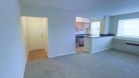 This single-floor unit presents an open-concept layout that seamlessly connects the main living area to the kitchen. The living space is finished with neutral wall-to-wall carpeting, lending a clean and understated aesthetic throughout. Natural light filters into the unit through a window visible in the kitchen area, contributing to a bright overall atmosphere.

The kitchen is partially open to the living area via a peninsula counter finished with a dark countertop surface, providing both a visual divider and an additional workspace or casual dining surface. The kitchen is equipped with stainless steel appliances, including a range, and features light wood cabinetry paired with a dark countertop. A sink is positioned beneath the window, allowing natural light while working in the kitchen. The cabinetry provides ample storage for everyday use.

An entryway with a doorway is visible to the left of the living area, offering a defined point of entry. The flooring near the entry transitions from carpet to hard surface, delineating zones within the space. The unit maintains a neutral color palette throughout, making it a versatile and move-in-ready environment.