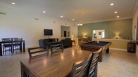 This single-floor space is designed for both leisure and social gatherings, offering a seamlessly connected open layout that encourages interaction and comfort throughout. The main area is anchored by a full-size pool table, making it an immediate focal point for recreational activity. Adjacent to it, a generous dining table with sturdy wooden chairs provides ample seating for group meals or casual gatherings. The lounge area features a dark sofa set arranged around a flat-screen television, creating a comfortable entertainment zone. Crown molding runs along the upper walls, lending a refined architectural detail to the space, while recessed lighting and a chandelier overhead provide layered illumination. The walls are finished in a soothing combination of neutral and sage green tones, contributing to a relaxed yet polished ambiance. An open archway leads to a secondary area furnished with accent chairs and additional décor, hinting at further flexible use of the space. Natural light filters in through windows visible in the background, brightening the interior and enhancing the overall sense of openness. The tile flooring throughout is practical and easy to maintain, complementing the warm wood tones of the furniture pieces. This space is well-suited for communal use, entertaining, or relaxed social activity.