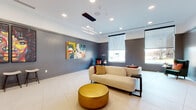Located on Dr. Martin Luther King Jr. Boulevard in Kansas City, Missouri, this single-floor community lounge space offers a thoughtfully designed environment that blends modern aesthetics with functional comfort. The open-plan layout is anchored by large-format porcelain tile flooring that stretches seamlessly throughout, lending the room an expansive, airy feel. Elevated ceilings with recessed lighting and a striking geometric linear pendant chandelier provide both ambient and accent illumination, enhancing the space's contemporary character. Floor-to-ceiling windows fitted with roller shades allow abundant natural light to filter in while offering views of the surrounding streetscape. The walls, finished in a refined grey tone with subtle ribbed paneling accents, serve as a sophisticated backdrop for a curated collection of bold, large-scale artwork—including a vibrant figurative diptych and colorful abstract compositions. Seating is arranged with a cream upholstered sofa, a deep green accent chair with vivid throw pillows, and a polished gold cylindrical ottoman, creating an inviting and visually dynamic gathering area. A built-in cabinetry wall with integrated task lighting and a utility sink provides practical functionality. Bar-height stools positioned near the art wall offer additional casual seating. The overall ambiance is modern, warm, and culturally expressive.