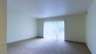 This single-floor space features a clean, minimalist layout centered around a spacious, open living area. The room is finished with wall-to-wall carpeting in a neutral beige tone, providing a soft, comfortable underfoot feel throughout. The walls are painted in a light, off-white hue, contributing to a bright and airy atmosphere.

A prominent feature of the living area is the large sliding glass door dressed with full-length vertical blinds, which span nearly the entire width of one wall. This door serves as the primary source of natural light, flooding the room with brightness and offering a view of the outdoors, where greenery is visible through the glass. The sliding door also provides direct outdoor access, adding both functionality and a sense of openness to the space.

The ceiling is smooth and finished in white, complementing the light palette of the room. A centrally placed ceiling vent suggests a climate control system is in place. The overall ambiance of the space is calm and neutral, presenting a versatile, move-in-ready environment well-suited to a variety of furnishing styles and uses.