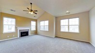 This single-floor home offers a welcoming and well-lit living area designed with comfort and functionality in mind. The main living room immediately impresses with its vaulted ceiling, which lends a sense of openness and grandeur to the space. A ceiling fan with integrated lighting is centrally mounted, providing both air circulation and ambient illumination. Soft, neutral-toned carpet covers the floor throughout, adding warmth underfoot and complementing the beige wall tones that create a calm, inviting atmosphere. A tile-surround fireplace with a classic white mantle serves as a natural focal point, ideal for relaxing evenings. Multiple large double-hung windows flood the room with natural light and offer views of the surrounding residential neighborhood, enhancing the sense of connection to the outdoors. Recessed lighting and standard ceiling vents are thoughtfully integrated into the ceiling, ensuring year-round comfort. The open layout provides flexible furniture arrangement options, making it well-suited for both everyday living and entertaining. The overall design balances traditional architectural details with a clean, understated aesthetic throughout.