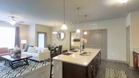This single-floor space showcases an open-concept layout that seamlessly integrates the kitchen, dining, and living areas into one cohesive environment. The kitchen serves as the centerpiece, anchored by a large island with light granite countertops, dark espresso cabinetry, and a stainless steel sink. Three pendant lights hang above the island, providing focused task lighting while contributing to the space's modern aesthetic. The flooring throughout is a warm hardwood tone, adding continuity and a polished feel across the main living areas.

The dining area sits adjacent to the kitchen, furnished with dark wood chairs and a matching table, complemented by a round mirror on the neutral-toned wall. The living room, visible beyond, features a white sofa atop a patterned area rug, with a window allowing natural light to filter in. A mirrored closet in the background provides a glimpse into a bedroom, suggesting a practical and connected floor plan. The overall color palette—neutral walls, dark wood accents, and light stone surfaces—creates a balanced, contemporary ambiance throughout the space.