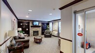 Located in Oakdale, PA, this well-appointed single-floor community space offers a thoughtfully designed layout that balances comfort and functionality across a generously sized footprint. The welcoming entrance hallway features recessed lighting, neutral carpeting, a resident mailbox wall, and decorative furnishings that set a warm, hospitable tone upon arrival. The lounge area is a standout, centered around a stone-accented fireplace flanked by rich dark wood built-in bookshelves and a wall-mounted television. Comfortable leather seating, a glass-top coffee table, and large windows dressed with draping curtains allow natural light to fill the room, creating an inviting atmosphere for relaxation and socializing. The expansive dining room is bright and open, illuminated by overhead fluorescent lighting and lined with large windows fitted with vertical blinds that bring in additional natural light. Numerous round tables with chairs provide generous seating capacity, supported by a service area and storage for utility carts. The kitchen is efficiently designed with warm wood cabinetry, granite-style countertops, stainless steel appliances including a refrigerator and dishwasher, and light hardwood-style flooring, making it well-suited for meal preparation and service. Together, these spaces create a functional and comfortable community environment.