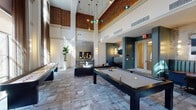 Located in Houston's 77008 zip code, Parlor is a thoughtfully designed communal gathering space that masterfully blends industrial character with contemporary elegance. The room commands attention with its dramatically soaring double-height ceilings, where exposed brick detailing meets crisp white walls, creating a striking architectural contrast. Large floor-to-ceiling windows line the perimeter, flooding the interior with abundant natural light while offering views of the surrounding greenery outside.

The main lounge area is anchored by a tufted leather sofa, a sleek coffee table, and a wall-mounted television set against a textured herringbone tile accent wall — perfect for relaxed social gatherings. A full-size pool table and a shuffleboard table provide engaging recreational options for residents and guests. Patterned tile-effect carpeting runs throughout, adding warmth and visual interest underfoot.

Tucked within the space is a semi-private nook featuring a channeled teal banquette, a round wooden table, and blue accent chairs — an intimate setting ideal for casual conversation or quiet work. Pendant lighting and wall sconces cast a warm, inviting glow throughout. Glass-panel French doors connect to an adjacent room, offering flexible use of the overall space. Lush indoor plants and curated artwork complete the upscale, welcoming atmosphere.
