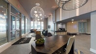 The Link's lobby is a thoughtfully designed, contemporary space that immediately communicates a sense of openness and sophistication. Soaring double-height ceilings anchor the room, drawing the eye upward to a striking collection of oversized spherical pendant lights with an intricate lattice design — a defining architectural feature that adds both drama and elegance.

Floor-to-ceiling glass windows line the perimeter, flooding the interior with abundant natural light and offering unobstructed views of the surrounding streetscape. The entrance is marked by a bold branded mat, setting a strong identity from the moment of arrival.

Large-format tile flooring in warm neutral tones extends throughout, grounding the space with a clean, polished finish. Multiple seating vignettes are thoughtfully arranged across the lobby, featuring a mix of upholstered lounge chairs in mustard yellow and teal tones, leather accent chairs, and modern round tables — each set atop layered, abstract area rugs that add color and texture.

A central reception desk with a dark, richly finished surface serves as the functional hub of the lobby, complemented by mailbox banks along the walls. A built-in coffee station and a wall-mounted display screen further enhance the space's utility. Wood-paneled wall accents and soft, recessed lighting contribute to a warm yet refined ambiance throughout.