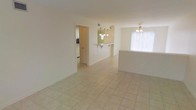 This two-story space features a bright, open main living area finished with light-toned ceramic tile flooring that extends throughout the ground floor, creating a cohesive and easy-to-maintain surface underfoot. The open layout connects the living area directly to a raised breakfast bar, which serves as a natural dividing point between the living and kitchen zones while maintaining an open, airy feel. The breakfast bar is topped with a tiled surface and is accented by a pendant chandelier overhead, adding a functional yet decorative focal point to the space. A ceiling fan is also present in the living area, supporting air circulation and comfort. The kitchen, visible through an adjoining doorway, features light-colored cabinetry and appears well-equipped for daily use. Walls throughout are finished in a neutral, warm white tone that reflects natural light effectively. A window fitted with vertical blinds allows natural light to filter into the living area. The overall design emphasizes clean lines, neutral finishes, and a practical open-concept flow between the primary living and kitchen spaces, offering a versatile foundation across both floors.