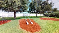 This outdoor recreational space is thoughtfully designed to provide a safe and engaging environment for play. The grounds are covered in well-kept green lawn, with multiple play structures strategically positioned throughout the area. A double swing set and a geodesic dome climber serve as the centerpiece attractions, each surrounded by vibrant reddish-brown rubber mulch beds that provide both a visually appealing contrast to the green grass and a protective safety surface underfoot. Additional play equipment, including what appears to be a climbing bar or similar structure, is also present on its own dedicated mulch pad. Mature oak trees are distributed across the space, their broad canopies offering generous natural shade — a welcome feature in what appears to be a warm, sunny climate. The entire area is enclosed by a tall white vinyl privacy fence that runs the perimeter, creating a secure and defined boundary. The open sky above and the lush tree cover combine to give the space a bright, airy, and natural ambiance, making it an inviting setting for active outdoor recreation.