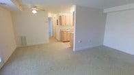 This single-floor space features an open-concept design that connects the living area to the kitchen, creating a functional and cohesive layout. The living area is finished with neutral wall-to-wall carpeting in a light beige tone, contributing to a warm and understated ambiance. A ceiling fan with a built-in light fixture is centrally mounted overhead, providing both ventilation and general illumination. The space is framed by clean white baseboards and painted walls in a soft, neutral gray-beige palette throughout.

The kitchen is partially separated from the living area by a half-wall partition, defining the space while maintaining an open feel. It features light wood cabinetry, both upper and lower, paired with a countertop that includes an integrated sink. The cabinetry offers ample storage, and the layout appears practical for everyday use. Overhead lighting in the kitchen ensures the workspace remains well-lit.

A hallway visible beyond the kitchen leads to additional areas of the unit, including what appears to be further rooms or utility spaces. The overall design is simple and functional, with consistent neutral tones and practical finishes throughout.