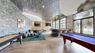 Located at 1920 Exchange Dr in Greenville, NC, this single-floor amenity lounge is a thoughtfully designed social space that seamlessly blends bold aesthetics with functional leisure areas. The interior is defined by striking textured wall coverings with an abstract, marble-like pattern in cool blue and grey tones, paired with a matching decorative ceiling treatment that adds dramatic visual depth throughout.

The space is anchored by a stunning central bar featuring an artistic, patterned facade with bar-height seating, illuminated by sculptural gold palm-leaf pendant lights that serve as a key design statement. Full-height glass garage doors line one wall, flooding the interior with natural light and offering expansive outdoor views of lush greenery and palm trees, creating a seamless indoor-outdoor ambiance.

Multiple lounge areas are thoughtfully arranged throughout the space, furnished with grey leather sofas, eclectic accent chairs, and tropical-patterned area rugs in rich forest green. Curated wall art, potted plants, and wood slat partition accents lend a warm, resort-inspired character to the interior.

A pool table positioned near the garage doors adds a recreational focal point, rounding out the space as a well-equipped social hub. Wood-look tile flooring ties all zones together, creating a cohesive and inviting atmosphere ideal for gathering and relaxation.